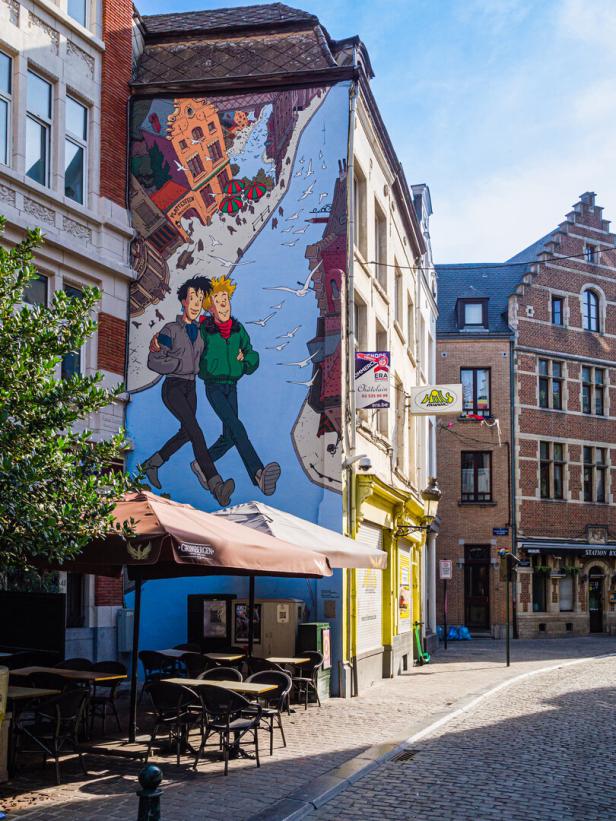 Straßenszene mit großem Comic-Wandbild an einer Hausfassade und Café-Tischen im Vordergrund.