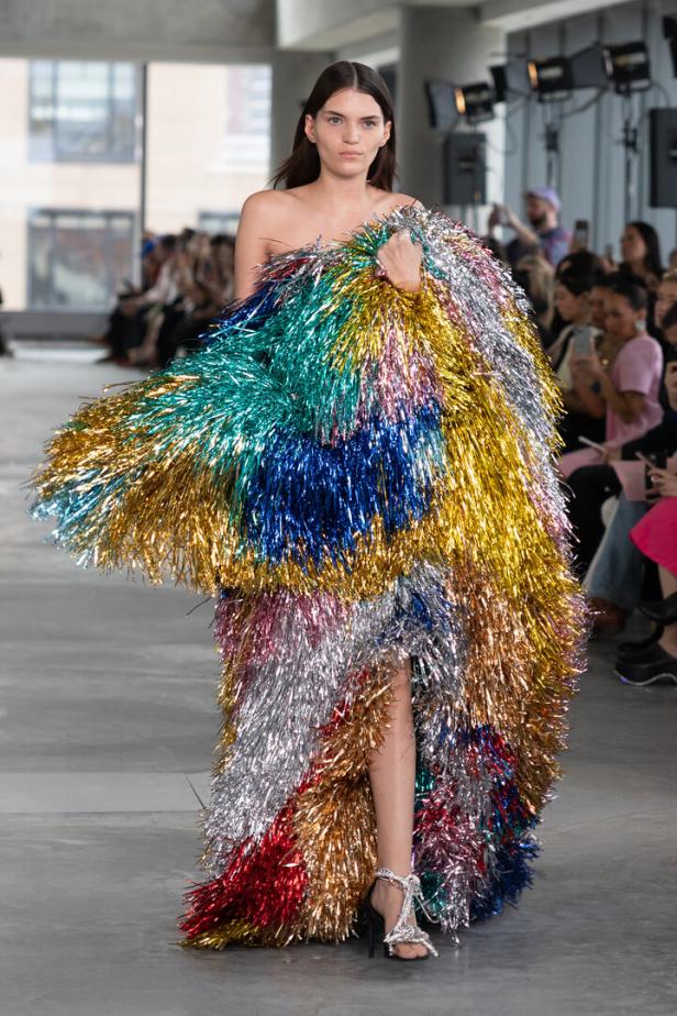 Model auf dem Laufsteg in einem bunten, glitzernden Fransen-Outfit vor sitzendem Publikum.