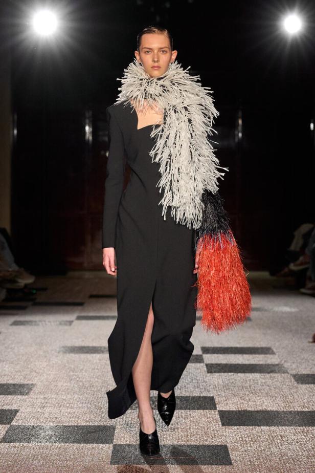 Model auf dem Laufsteg in schwarzem Kleid mit auffälligem weiß-rotem Fransenschal.