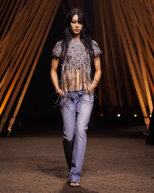 Model auf dem Laufsteg in Fransenoberteil und Jeans, zwischen beleuchteten Holzstreben.