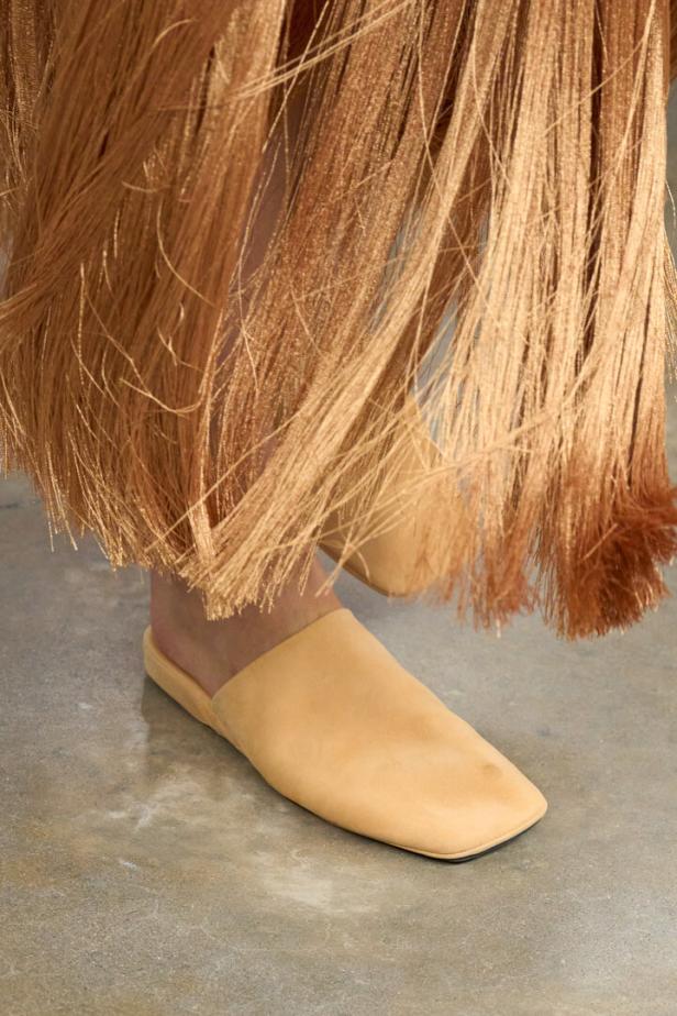 Beiger flacher Schuh unter einem langen Fransensaum auf grauem Boden.