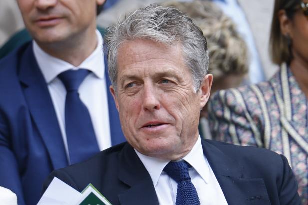 Der Schauspieler Hugh Grant trägt einen dunklen Anzug mit Krawatte und blickt aufmerksam zur Seite.