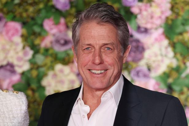 Hugh Grant lächelt vor einem Hintergrund aus Blumen.
