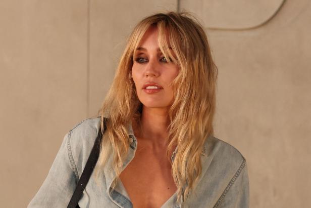 Miley Cyrus mit blondem, gestuftem Haar und einem hellen Jeanshemd vor einer beigen Wand.