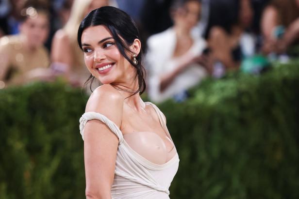 Kendall Jenner lächelt in einem hellen, schulterfreien Kleid vor einem unscharfen Hintergrund in die Kamera.