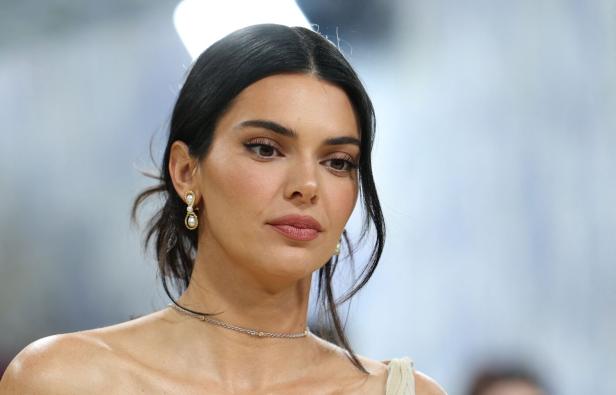 Kendall Jenner mit dunklem Haar, Perlenohrringen und einer feinen Halskette vor einem hellen Hintergrund.