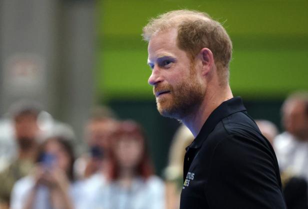 Prinz Harry trägt ein schwarzes Poloshirt mit dem Logo der Invictus Games und blickt aufmerksam zur Seite.