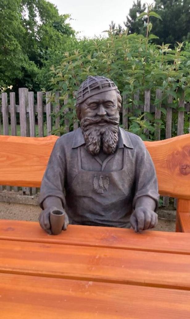 Holzfigur eines bärtigen Mannes mit Hut sitzt an einem Tisch und hält einen Becher.
