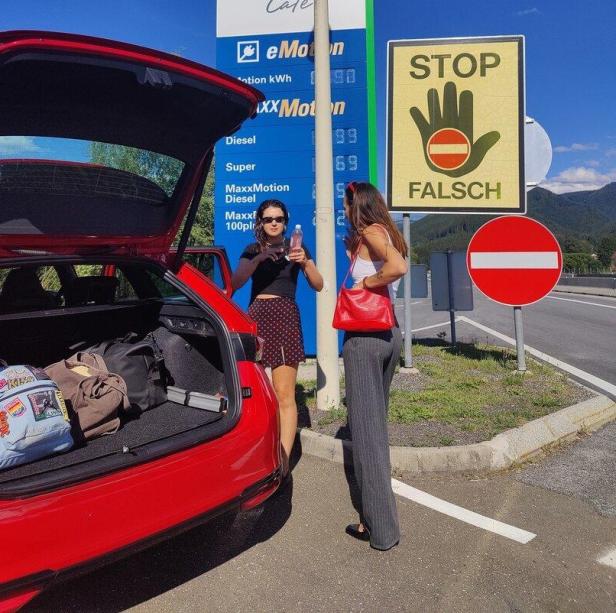 Zwei Frauen stehen neben einem roten Auto an einer Tankstelle mit Verbotsschildern und Preisanzeige.