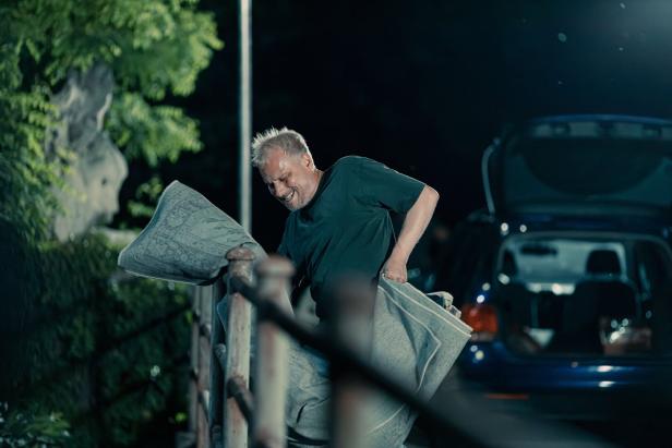 Ein Mann mit grauem Haar schüttelt nachts einen Teppich über einem Metallgeländer aus, während im Hintergrund ein Auto mit geöffnetem Kofferraum steht.