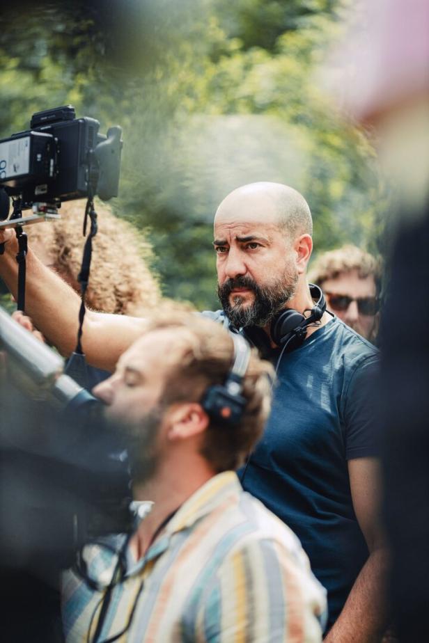 Bärtiger Mann mit Kopfhörern bedient eine Filmkamera im Freien, umgeben von Crewmitgliedern.