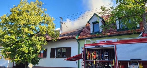 Feuerwehr löscht Rauch aus dem Dach eines Wohnhauses, daneben ein Einsatzfahrzeug mit offener Ausrüstung.