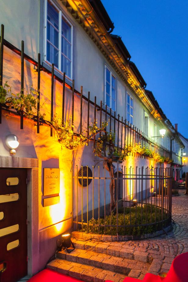Abendliche Gasse mit beleuchtetem Altbau, Schmiedeeisenzaun, Treppe und blauen Fenstern.