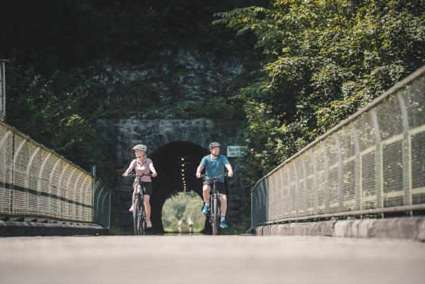 Zwei Radfahrende fahren auf einer Brücke vor einem dunklen Tunnel durch grünes Waldgebiet.