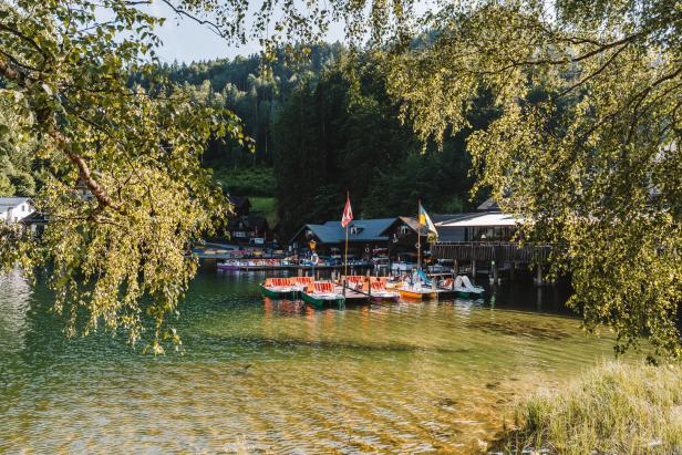 Bunte Tretboote liegen an einem Steg vor einem Ufer mit Häusern an einem sonnigen See.