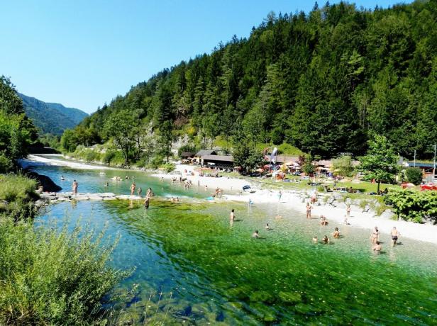 Menschen baden in einem klaren grünblauen Fluss zwischen Waldhängen und Kiesufern an einem sonnigen Sommertag.