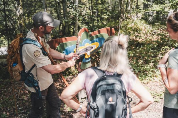 Ein Führer des Wildnisgebiets erklärt zwei Frauen im Wald an einer bunten Holztafel in Vogelform die Bedeutung einer Feder.