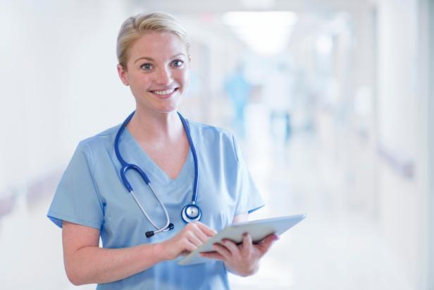 Eine lächelnde Pflegekraft in blauer Dienstkleidung mit Stethoskop hält ein Tablet im Krankenhaus.