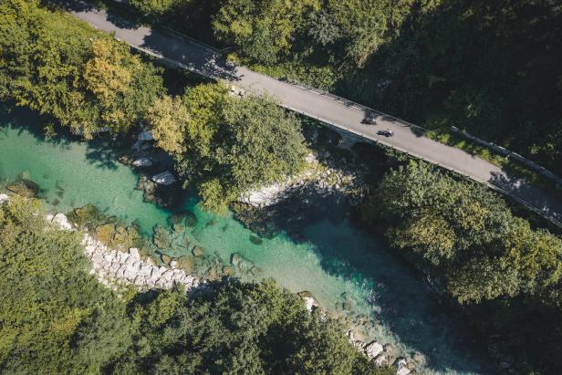 Luftaufnahme eines Gebirgsbachs mit Brücke und zwei Radfahrern zwischen dichtem Wald.