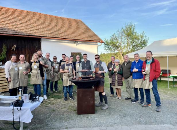 Gruppe von Menschen mit Schürzen steht vor einem Grill bei einer Gartenfeier neben einem Haus.