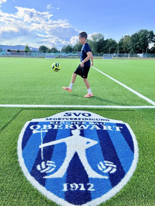 Junge jongliert einen Fußball auf einem Kunstrasenplatz mit Vereinslogo.