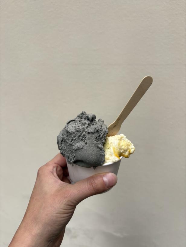 Hand hält einen Becher mit schwarzem und weißem Eis samt Holzlöffel vor einer hellen Wand.