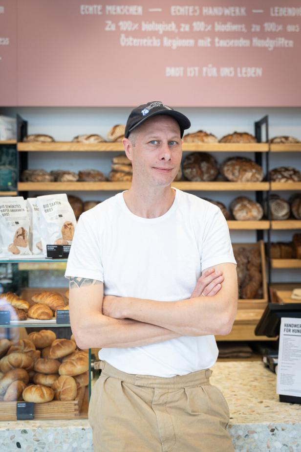 Mann mit weißem T-Shirt und Kappe steht in einer Bäckerei vor Regalen mit Brot und Gebäck.