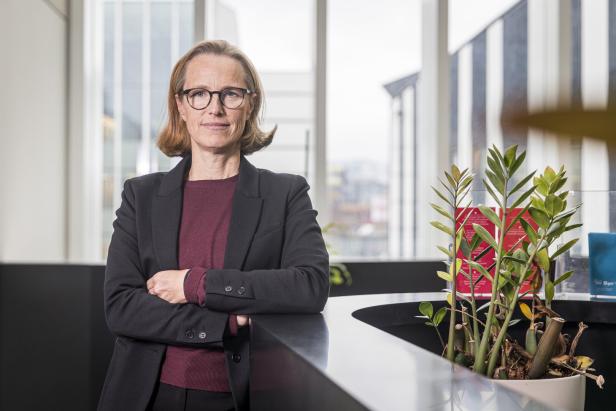 Eine Frau mit Brille und schwarzem Blazer steht mit verschränkten Armen in einem hellen Büro neben einer Pflanze.