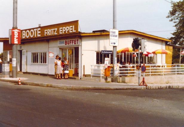 Straßenecke mit Imbiss „Boote Fritz Eppel“, Strandrestaurant, Schildern und einigen Menschen vor dem Eingang.
