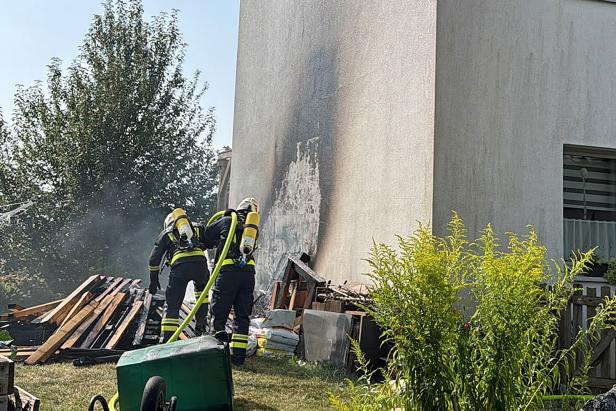 Feuerwehrleute löschen einen Brand an einer Hauswand neben Holzstapeln und Rauch.