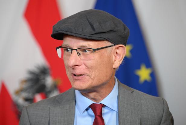 Ein Mann mit schwarzer Schiebermütze, Brille und Anzug spricht vor österreichischen und EU-Flaggen.
