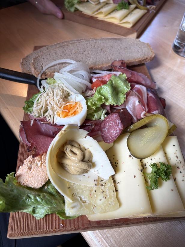 Rustikale Brotzeitplatte mit Käse, Wurst, Ei, Salat, Zwiebeln und Gurken auf Holzbrett.