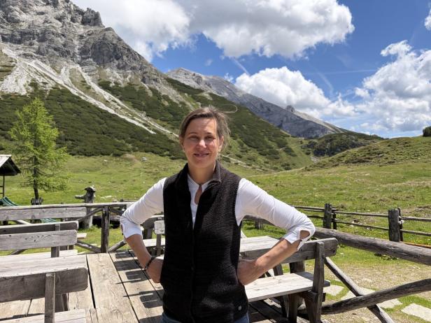 Frau steht lächelnd vor einer Berglandschaft mit Wiesen, Felsen und blauem Himmel.