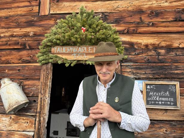 Ein Mann in Trachtenweste und Hut steht vor der Holzhütte „Tauernkarle“ mit einem „Herzlich willkommen“-Schild.