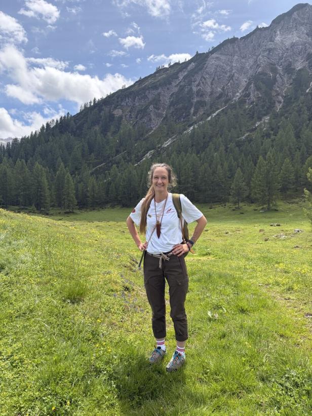 Eine Frau steht in einer Almwiese vor bewaldetem Bergmassiv unter blauem Himmel.