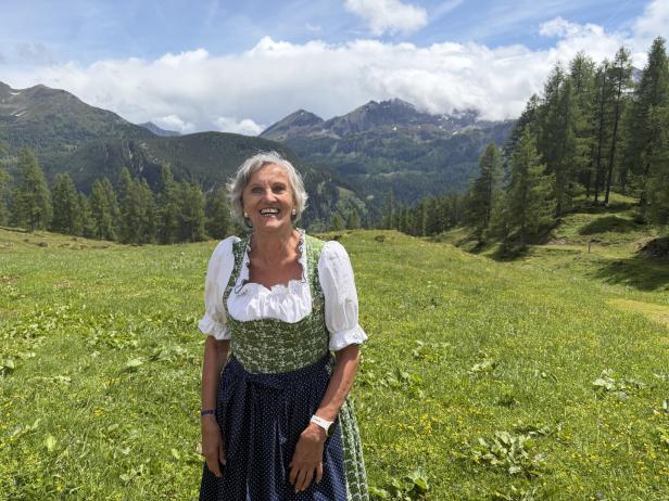 Eine lächelnde Frau im Dirndl steht auf einer grünen Bergwiese vor einer alpinen Kulisse.