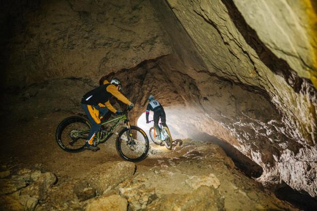 Zwei Mountainbiker fahren mit Helmlampen durch eine enge Felsgrotte bergab.