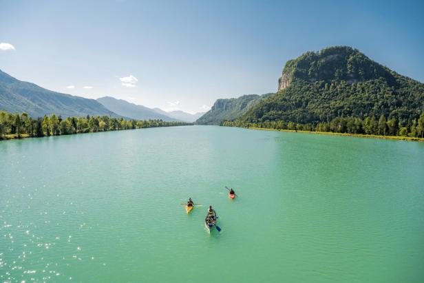 Drei Boote mit Menschen paddeln auf einem türkisfarbenen Fluss vor einer bewaldeten Bergkulisse.