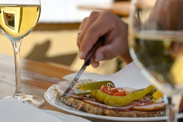 Weißwein und Jause auf einem Tisch, eine Hand schneidet Brot mit Aufschnitt.