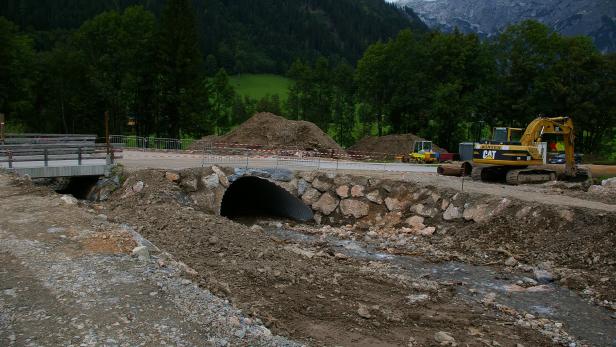 Baustelle mit Bagger an einem Bach unter einer Brücke im Gebirge.