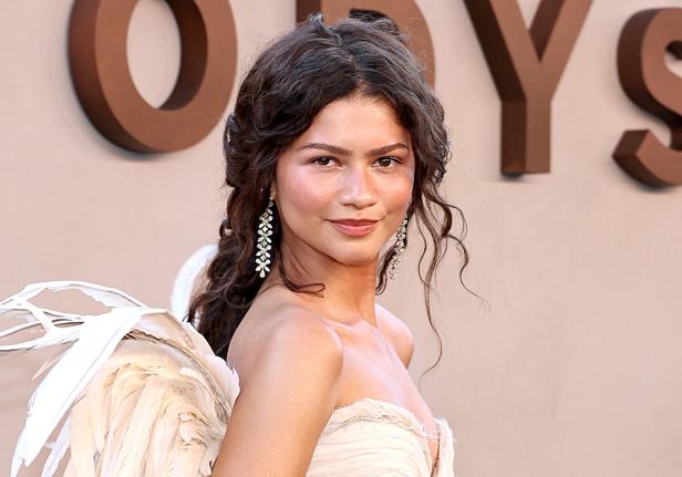 Zendaya lächelt in einem hellen Kleid mit Federdetails und trägt auffällige Ohrringe vor einem beigen Hintergrund.