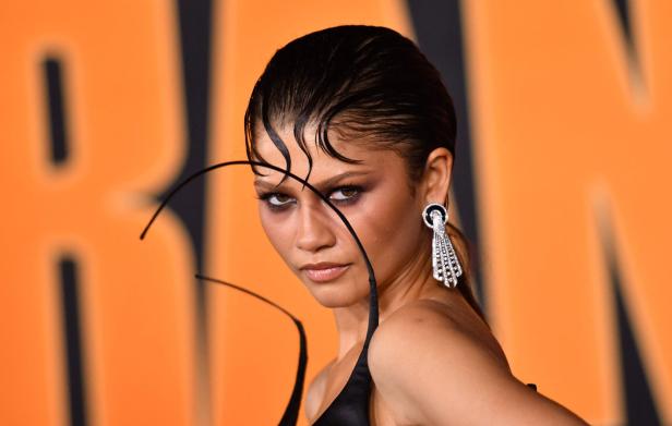 Zendaya trägt eine elegante Hochsteckfrisur mit markanten, ins Gesicht fallenden Haarsträhnen und auffälligen Ohrringen vor einem orangefarbenen Hintergrund.