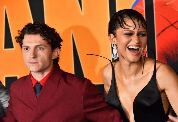 Tom Holland und Zendaya lachen gemeinsam vor einem orangefarbenen Hintergrund.