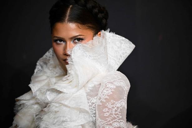 Zendaya trägt ein weißes Spitzenoberteil mit hohem Kragen vor einem schwarzen Hintergrund.