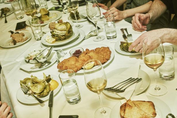 Menschen sitzen im Salzamt an einem gedeckten Tisch mit Wiener Schnitzel, Artischocken, Weißwein und Wassergläsern.