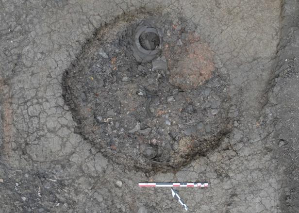Archäologische Grabung mit dunkler, kreisförmiger Verfärbung und Keramik- bzw. Gefäßresten im Boden.