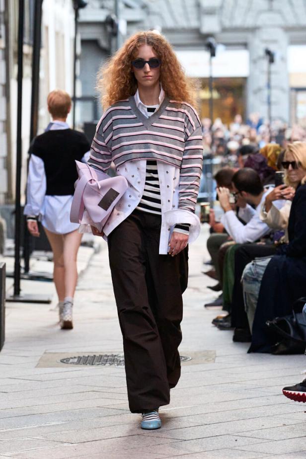 Model auf dem Laufsteg mit lockigem Haar, Sonnenbrille, gestreiftem Layer-Look und lilafarbener Tasche vor sitzendem Publikum.