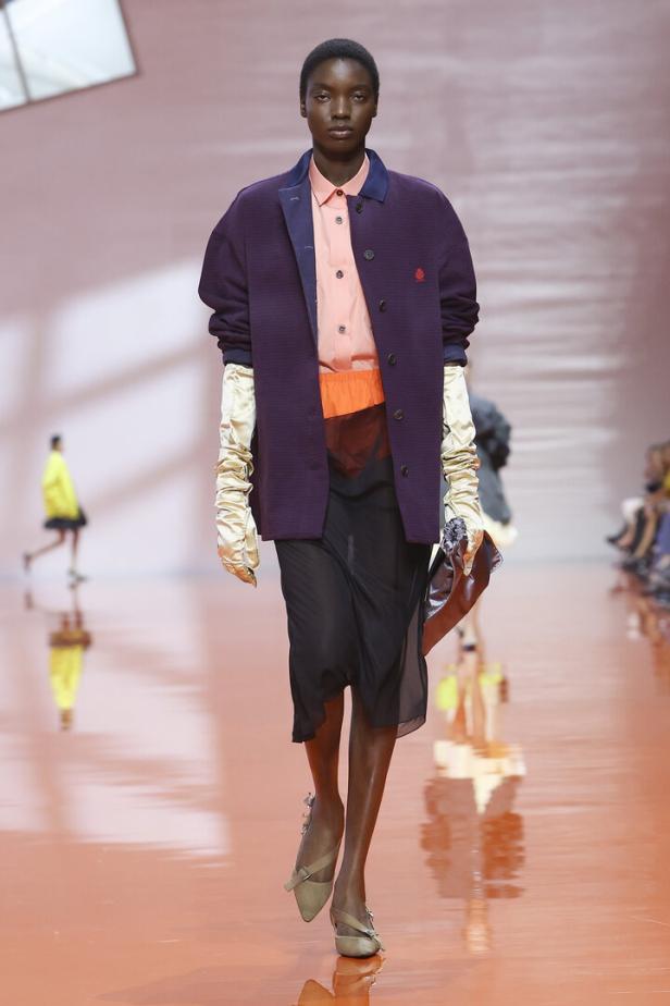 Ein Model präsentiert auf einem Laufsteg eine dunkle Jacke über einem pfirsichfarbenen Hemd, kombiniert mit einem schwarzen Rock und langen Handschuhen.
