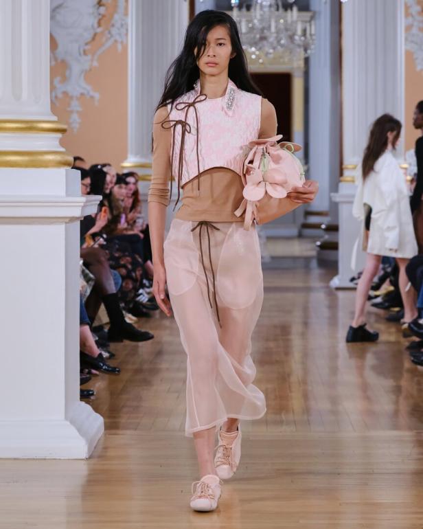 Model läuft auf dem Laufsteg in zartrosa Outfit mit transparenter Hose und hält eine rosa Tasche.