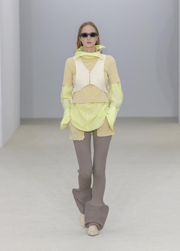 Ein Model präsentiert auf einem Laufsteg ein durchsichtiges gelbes Oberteil mit weißer Weste, graue gerippte Hosen mit weitem Saum und eine dunkle Sonnenbrille.